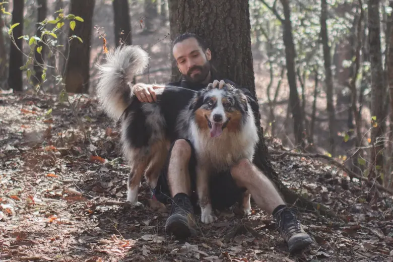 Julio sitting against a tree in the woods with two Australian Shepherds, one of them panting happily