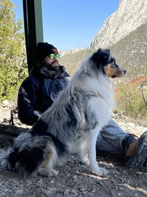 Julio sitting in the shade in the mountains with Thor, his merle Australian Shepherd, beside him